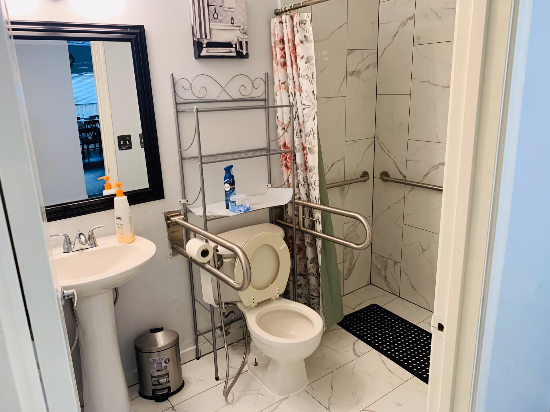 A clean and accessible bathroom featuring a white pedestal sink with soap dispenser, a toilet with safety rails on both sides, a metal shelving unit above the toilet holding cleaning supplies, and a walk-in shower with marble-patterned tiles and grab bars. A floral shower curtain is partially drawn to the side.
