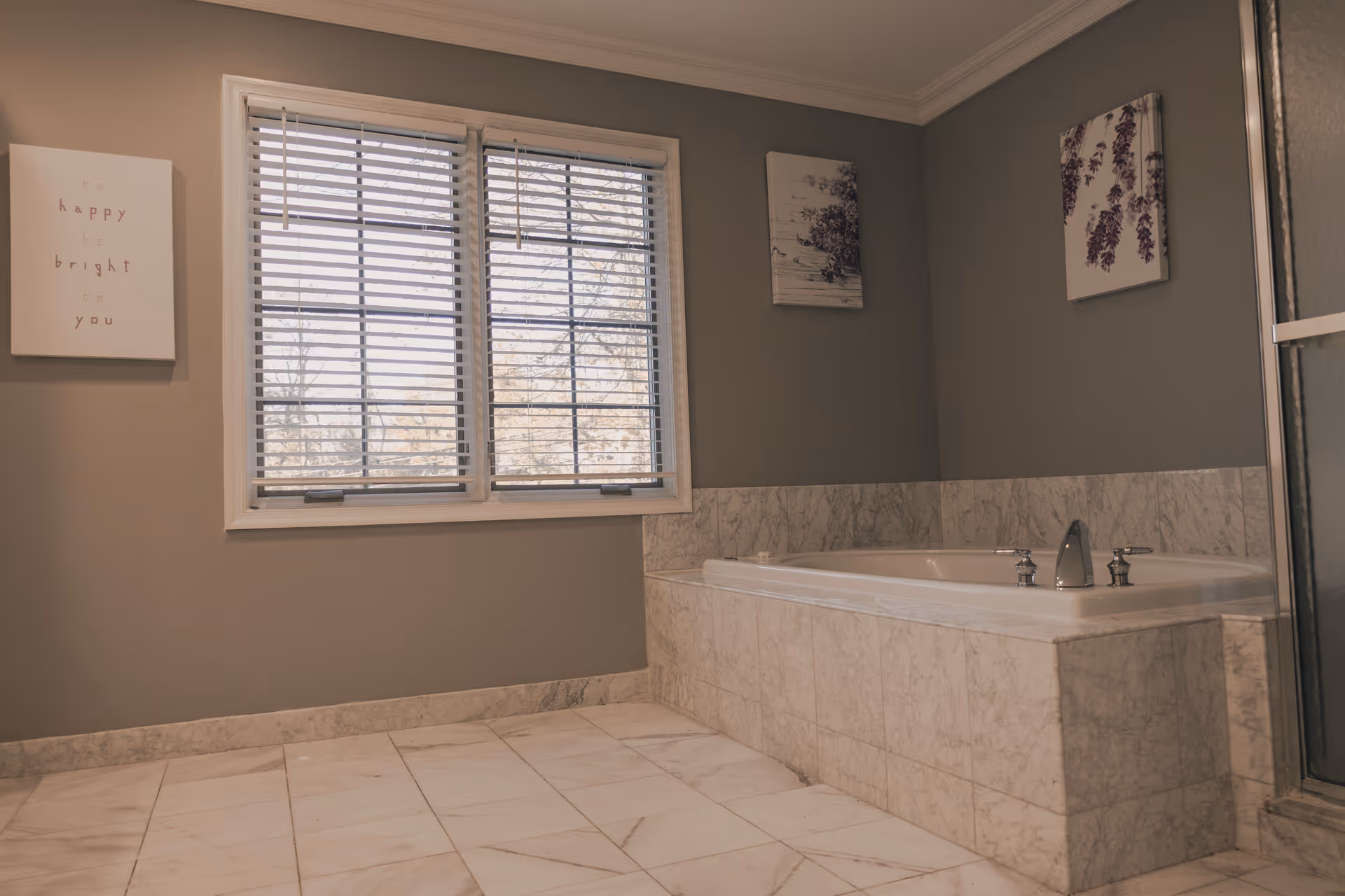A bathroom with a large built-in bathtub surrounded by marble tiles. There is a window with blinds above the tub, and three pieces of wall art hang on the walls. The floor is also tiled in marble.