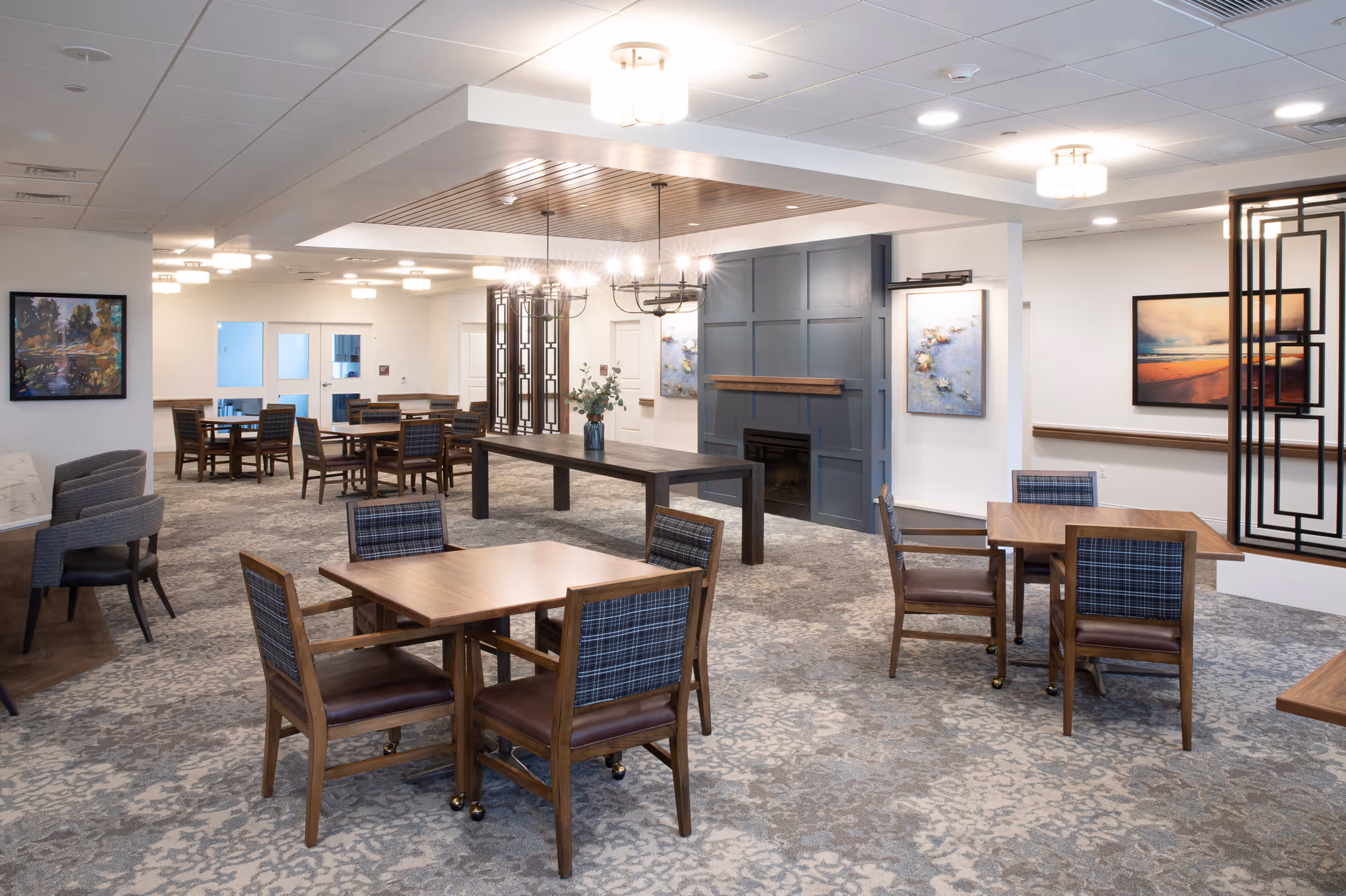Spacious, well-lit communal dining and activity room with multiple wooden tables and chairs, decorative lighting, and artwork on the walls.