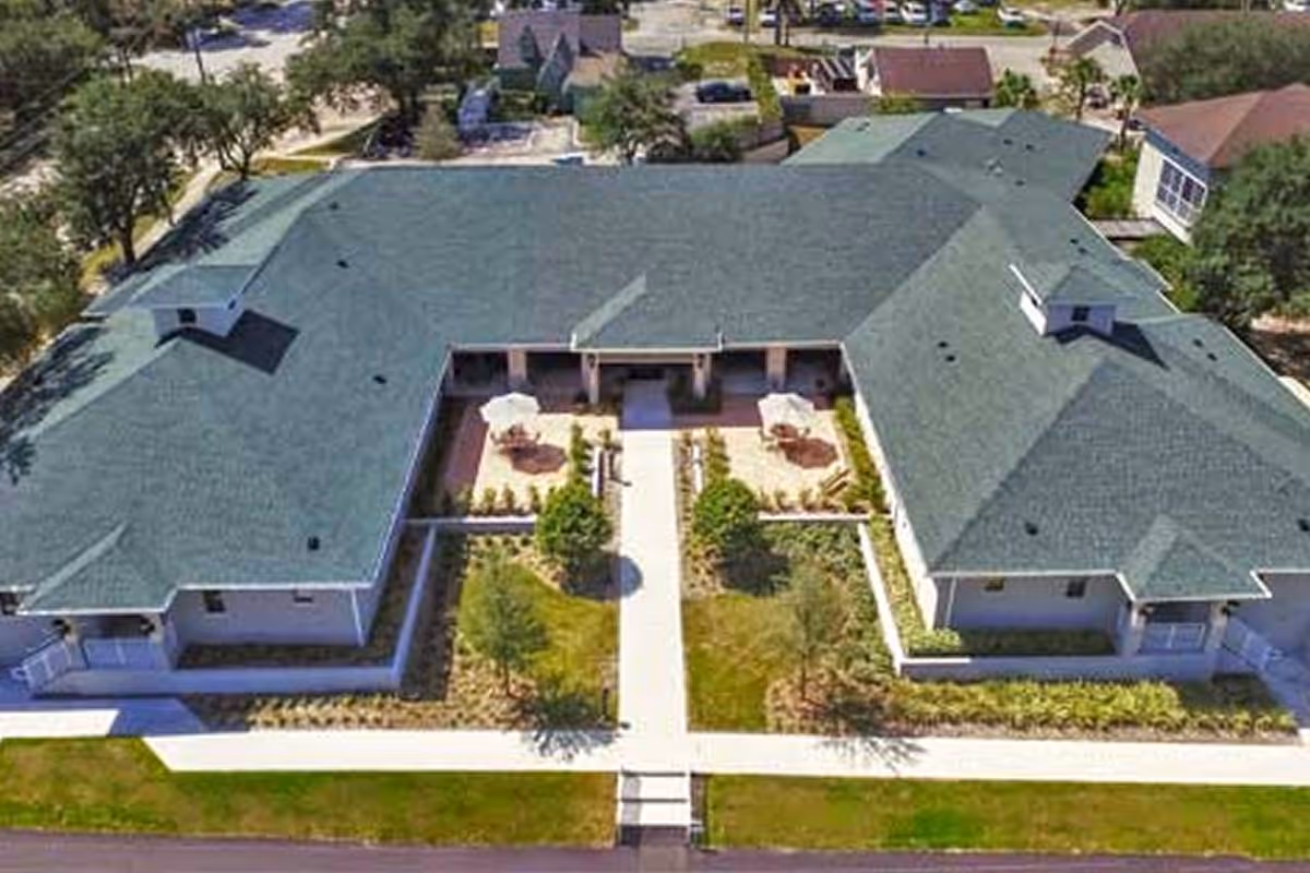 Aerial view of a single-story building with a green roof surrounding a central courtyard featuring two patio areas with umbrellas and seating, landscaped with trees and shrubs, and a walkway leading to the entrance.