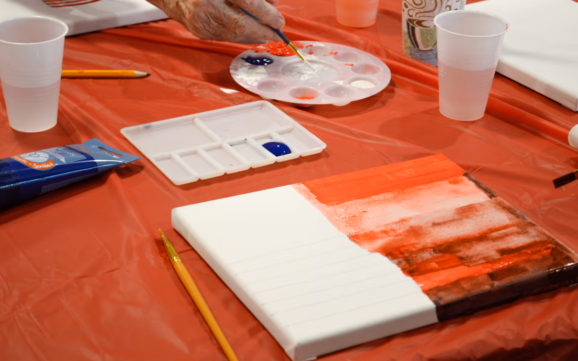 A close-up view of a painting activity setup on a red plastic-covered table, featuring a canvas partially painted with red and brown colors, paintbrushes, a paint palette with various colors, a tube of blue paint, and plastic cups. A hand is seen holding a paintbrush applying white paint on the palette.