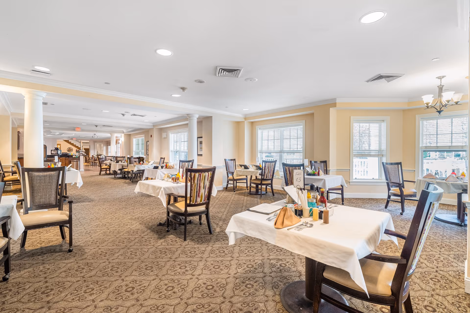 A bright and spacious dining room in a senior living facility with multiple tables covered with white tablecloths, set with condiments and napkins. The room features large windows allowing natural light to fill the space, beige walls, carpeted floors, and several wooden chairs arranged around the tables.