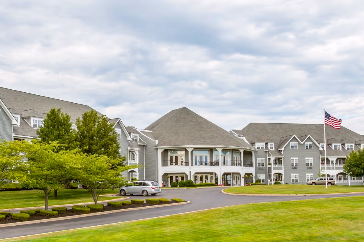Exterior view of a large senior living facility building with gray siding and multiple windows. The building has a central section with a covered balcony and a circular driveway in front. There are green lawns, trees, and an American flag on a flagpole near the entrance under a cloudy sky.