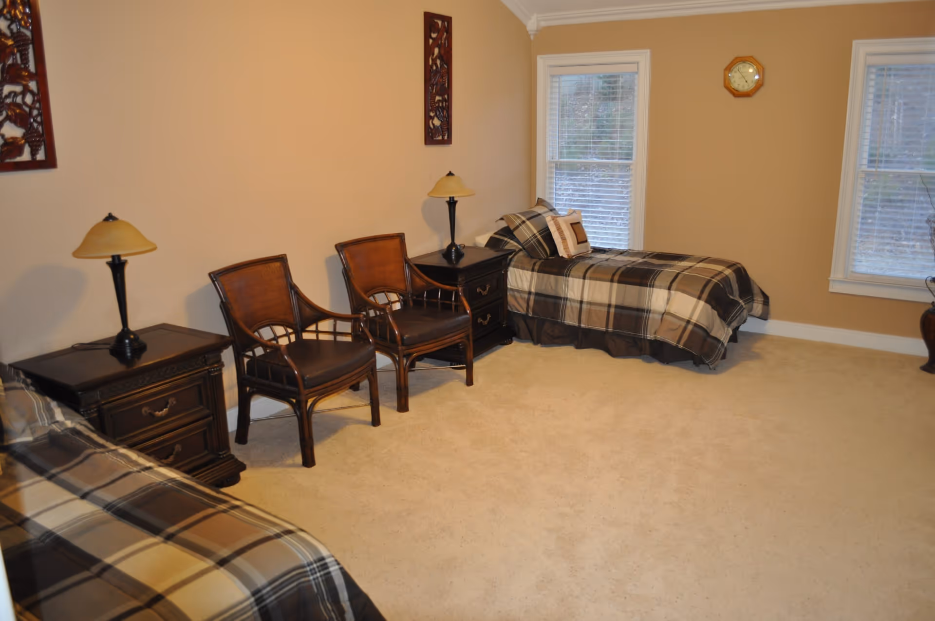 A senior living bedroom with two single beds covered in plaid bedding, two wooden chairs with dark cushions, two dark wooden nightstands each with a lamp, beige walls, two windows with white blinds, and a wall clock between the windows.