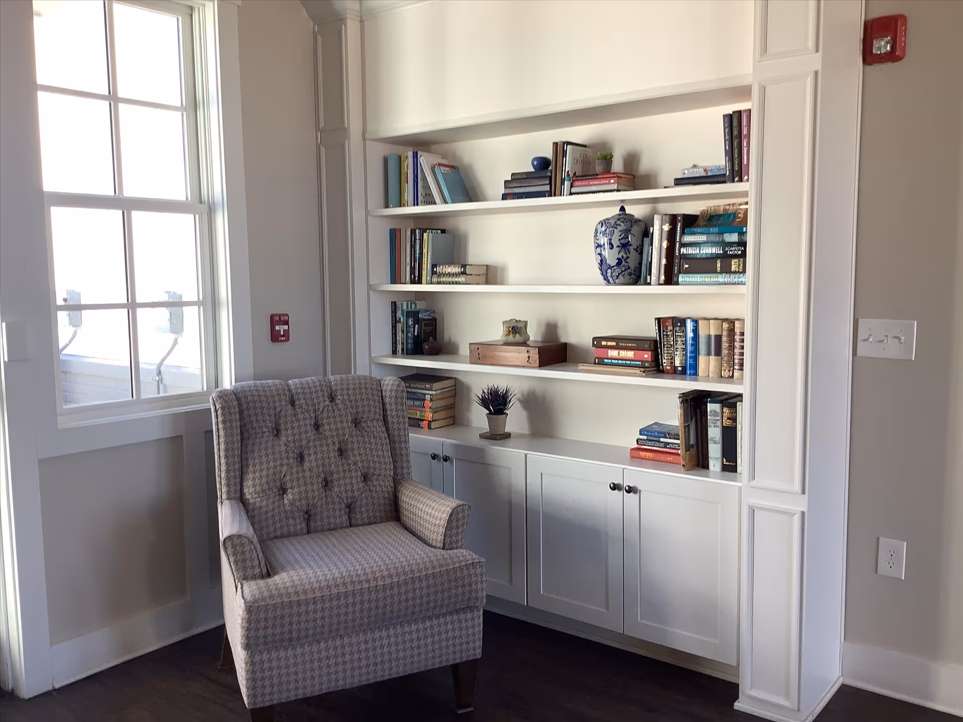 A cozy corner of a room featuring a patterned upholstered armchair next to a large window with white trim. Behind the chair is a built-in white bookshelf filled with various books and decorative items, including a blue and white vase and small potted plants. The room has light-colored walls and dark wood flooring.