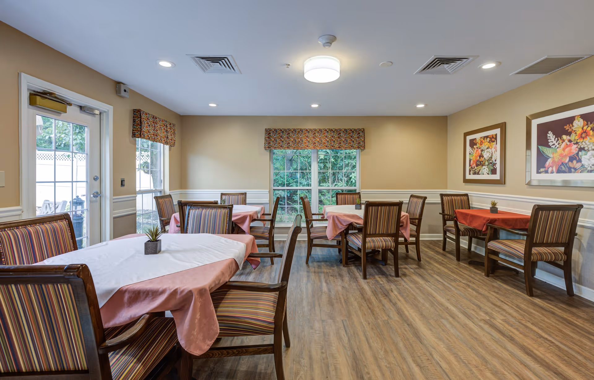 A dining room with several wooden tables covered with pink and white tablecloths, surrounded by wooden chairs with striped cushions. The room has beige walls with white wainscoting, two framed floral paintings on the right wall, and windows with patterned valances letting in natural light. There is a door on the left side leading outside, and the floor is wood laminate.