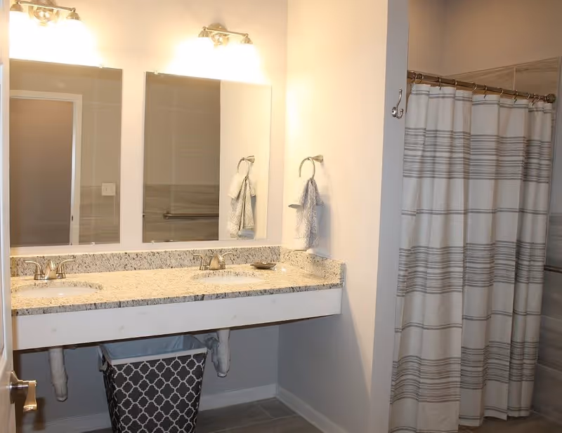 A bathroom with a double sink vanity featuring granite countertops, two mirrors above the sinks, two wall-mounted light fixtures, a towel ring with a hand towel, and a shower area with a striped shower curtain.