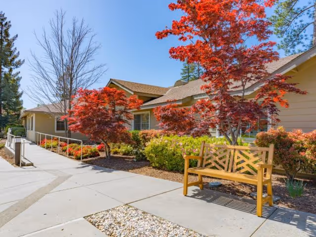 Outdoor view of a senior living facility with a paved walkway, a wooden bench, and vibrant red and green foliage. The building has beige siding and a sloped roof under a clear blue sky.