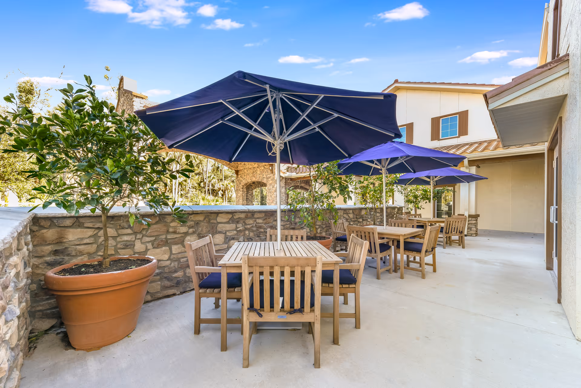Outdoor patio area at Tuscan Gardens of Palm Coast Senior Living with wooden tables and chairs, each shaded by large blue umbrellas. There are potted plants along a stone wall, and the building exterior is visible under a clear blue sky.