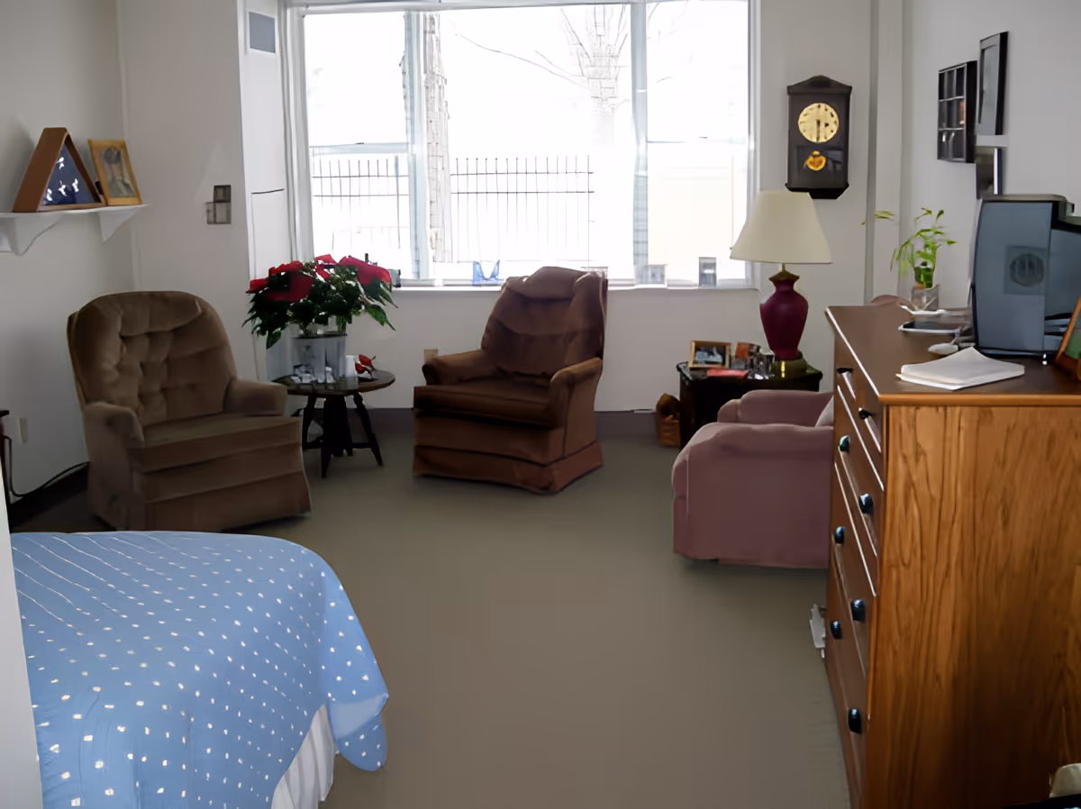 A cozy living room area with three upholstered armchairs, a small round table with a poinsettia plant, a wooden dresser with a lamp and TV on top, a wall clock, and a large window letting in natural light.