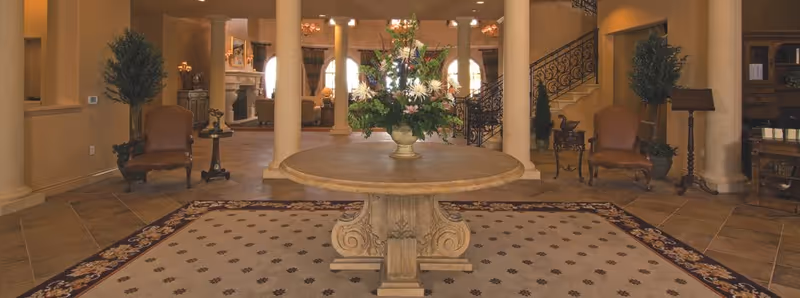 Wide entry lobby with a round table topped by a large floral arrangement, flanked by columns, seating, and a staircase.