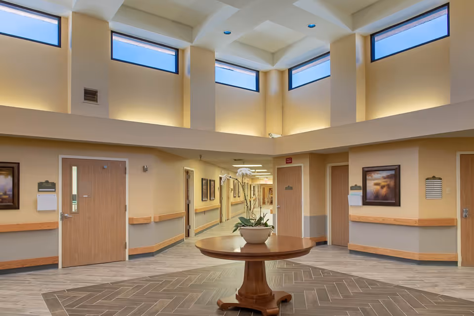 A spacious hallway in a senior living facility with high ceilings and several tall windows near the ceiling allowing natural light to enter. The walls are painted beige with a gray lower section and wooden handrails. There are several closed wooden doors along the hallway, framed artwork on the walls, and a round wooden table with a potted plant in the center of the floor.