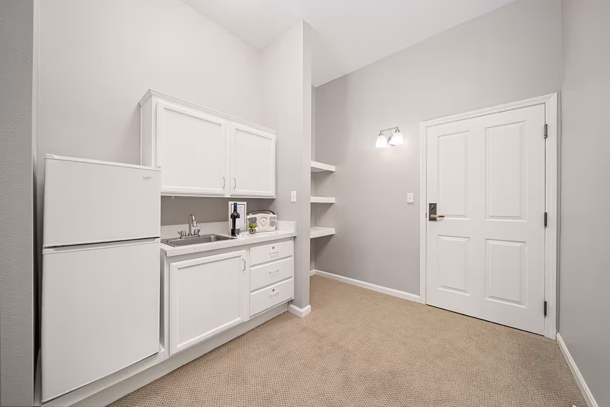 Small kitchenette with white cabinets, a sink, mini refrigerator, open shelving, and a closed door in a neutral-toned room.
