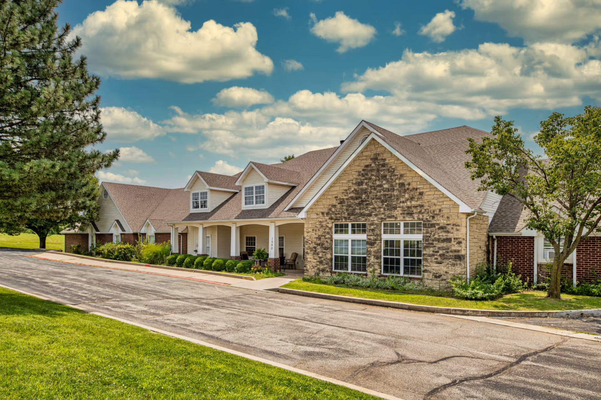 Exterior view of Creekside Village senior living facility showing a large building with a combination of brick and stone facade, multiple windows, and a covered porch area. The building is surrounded by green grass, trees, and a paved driveway under a partly cloudy sky.