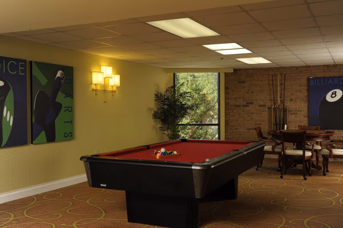 A recreational room with a red felt pool table in the center, a round wooden table with chairs in the background, billiard cues standing against a brick wall, decorative wall art featuring dice and billiards, a large window with a view of greenery, and warm lighting from wall sconces and ceiling lights.