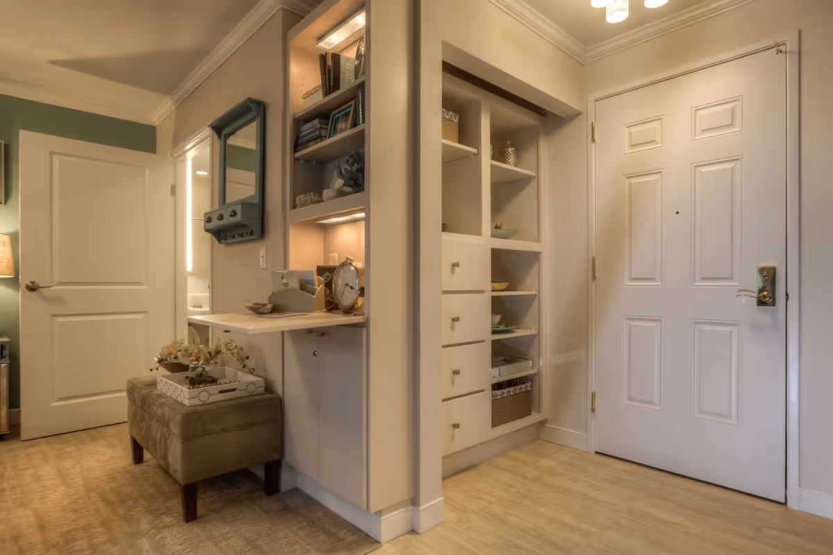 Interior view of an entryway area in a senior living facility with a white front door, built-in shelves with decorative items, a small vanity desk with a clock and decor, a green cushioned bench, and a partially open door leading to another room.