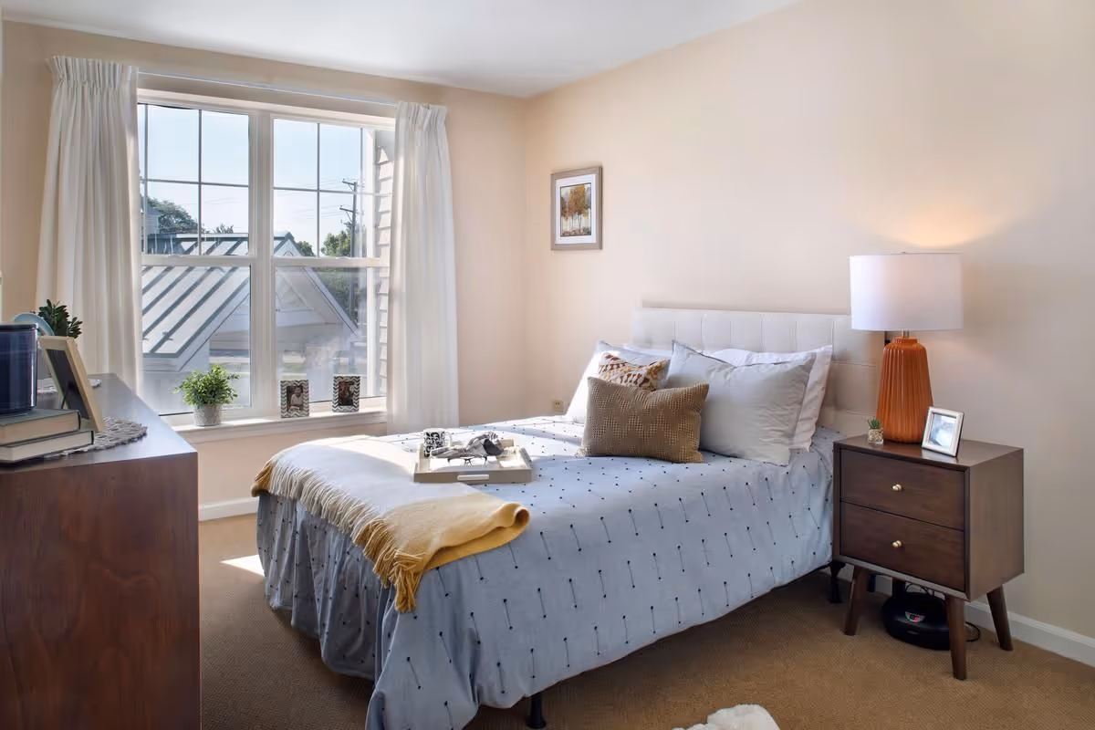 Sunlit bedroom with a bed dressed in light linens, a wooden nightstand with a lamp, a dresser, and a large window overlooking nearby rooftops.