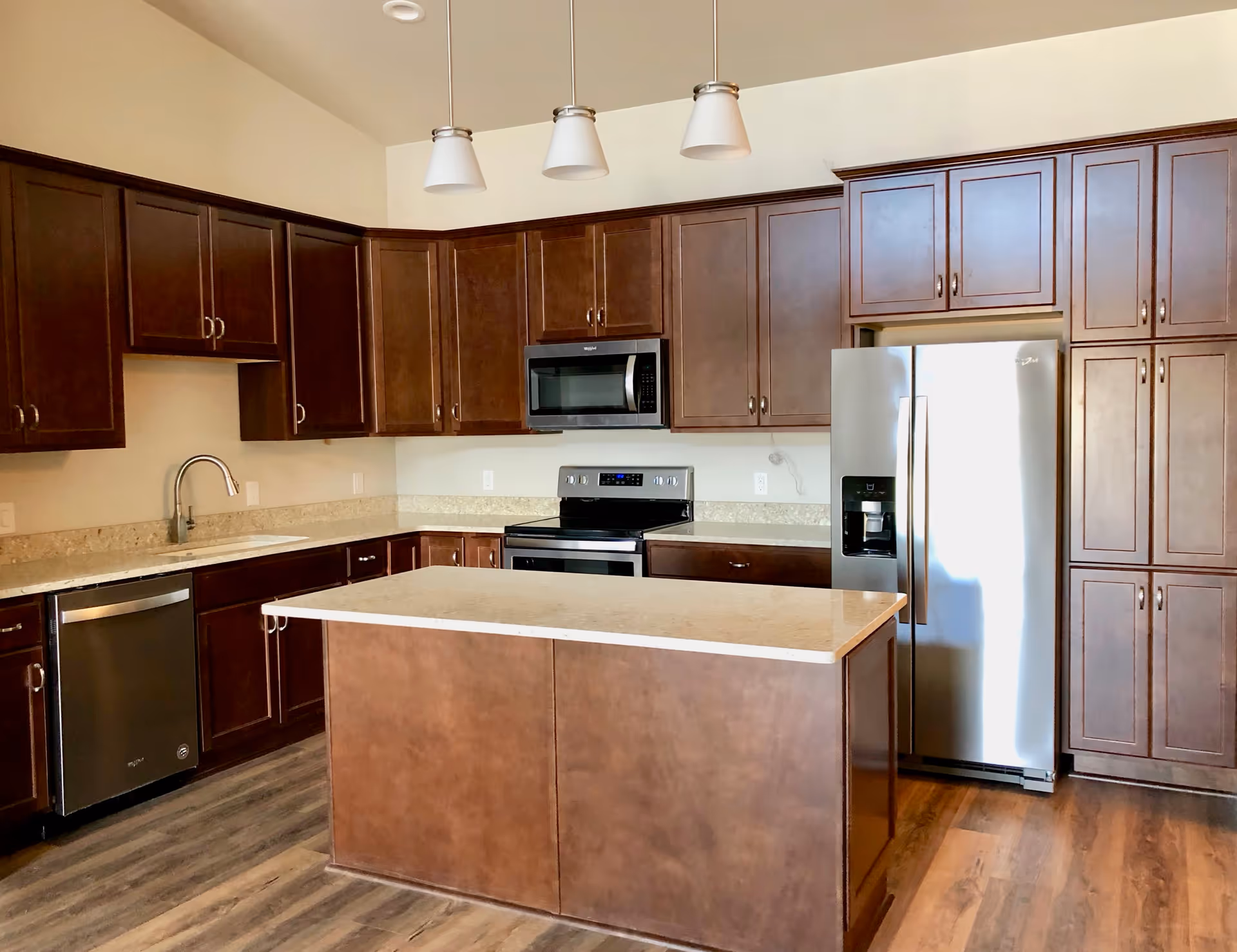 Modern kitchen with dark wood cabinets, a central island with a light countertop, stainless steel appliances including a refrigerator, microwave, stove, and dishwasher, and three pendant lights hanging from the ceiling.