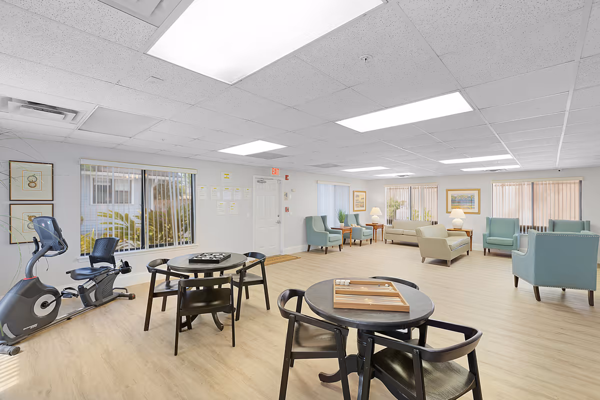 A spacious senior living common area with light wood flooring and white walls. The room features several seating areas with light blue and beige armchairs and sofas, side tables with lamps, and framed artwork on the walls. There are two round black tables with chairs, one with a backgammon game and the other with a tic-tac-toe game. A stationary exercise bike is positioned near a window with vertical blinds. The ceiling has recessed lighting panels, and there is an exit door on one wall.
