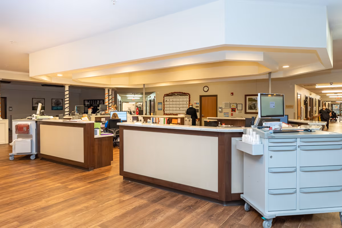 Interior view of a nursing and rehabilitation center's nurse station area with multiple desks, computer monitors, and staff working. The space has wood flooring, white walls, and a ceiling with recessed lighting. Hallways extend from the nurse station, and there are various framed pictures and a clock on the walls.