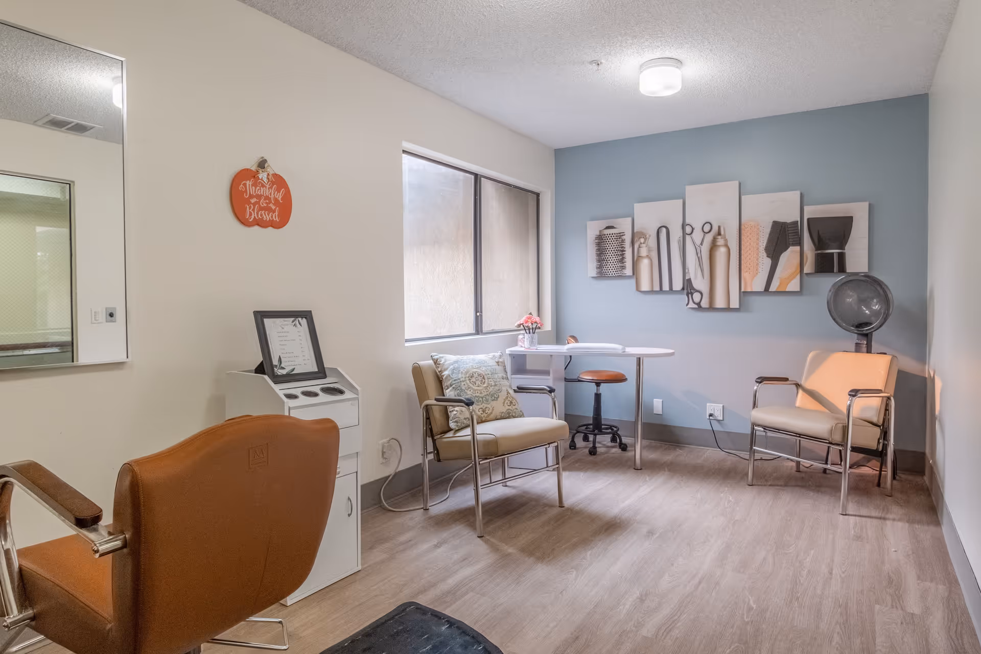 A small salon room with two beige salon chairs, a brown salon chair, a hair dryer, a small white table with a brown stool, and wall art depicting hair styling tools. The room has light wood flooring, a large window, and a wall painted light blue.