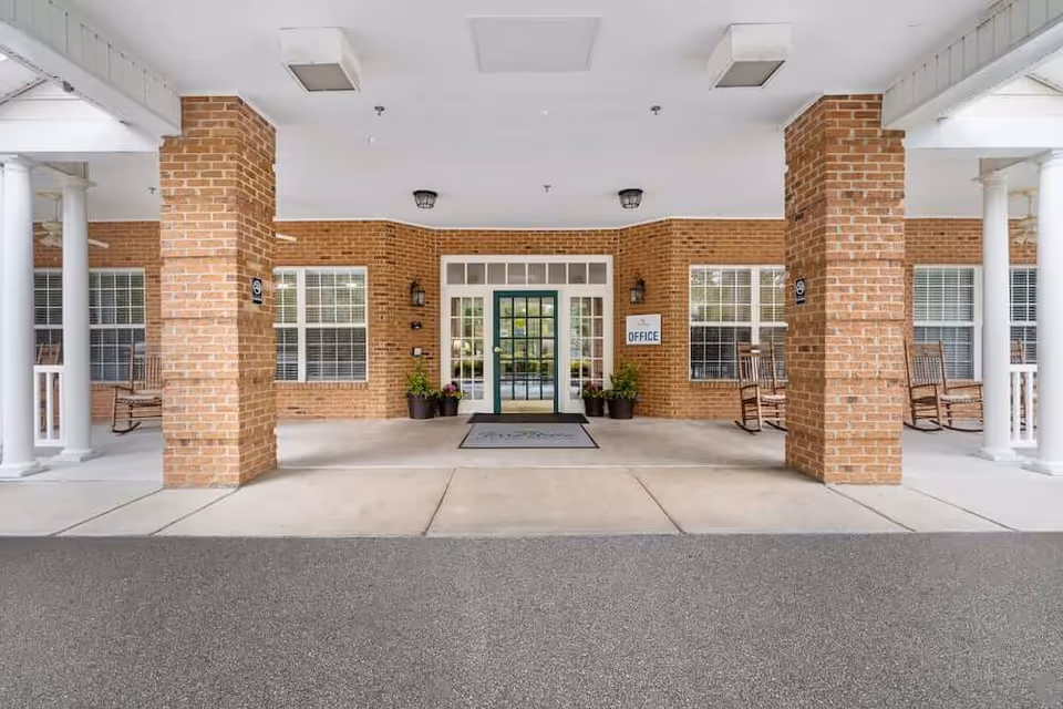 Entrance area of a brick building with two large brick columns and white pillars supporting a covered walkway. There are rocking chairs on either side near windows with white blinds. A glass door with sidelights and a transom window above leads into the office, indicated by a sign on the right side of the door. Potted plants are placed near the entrance.
