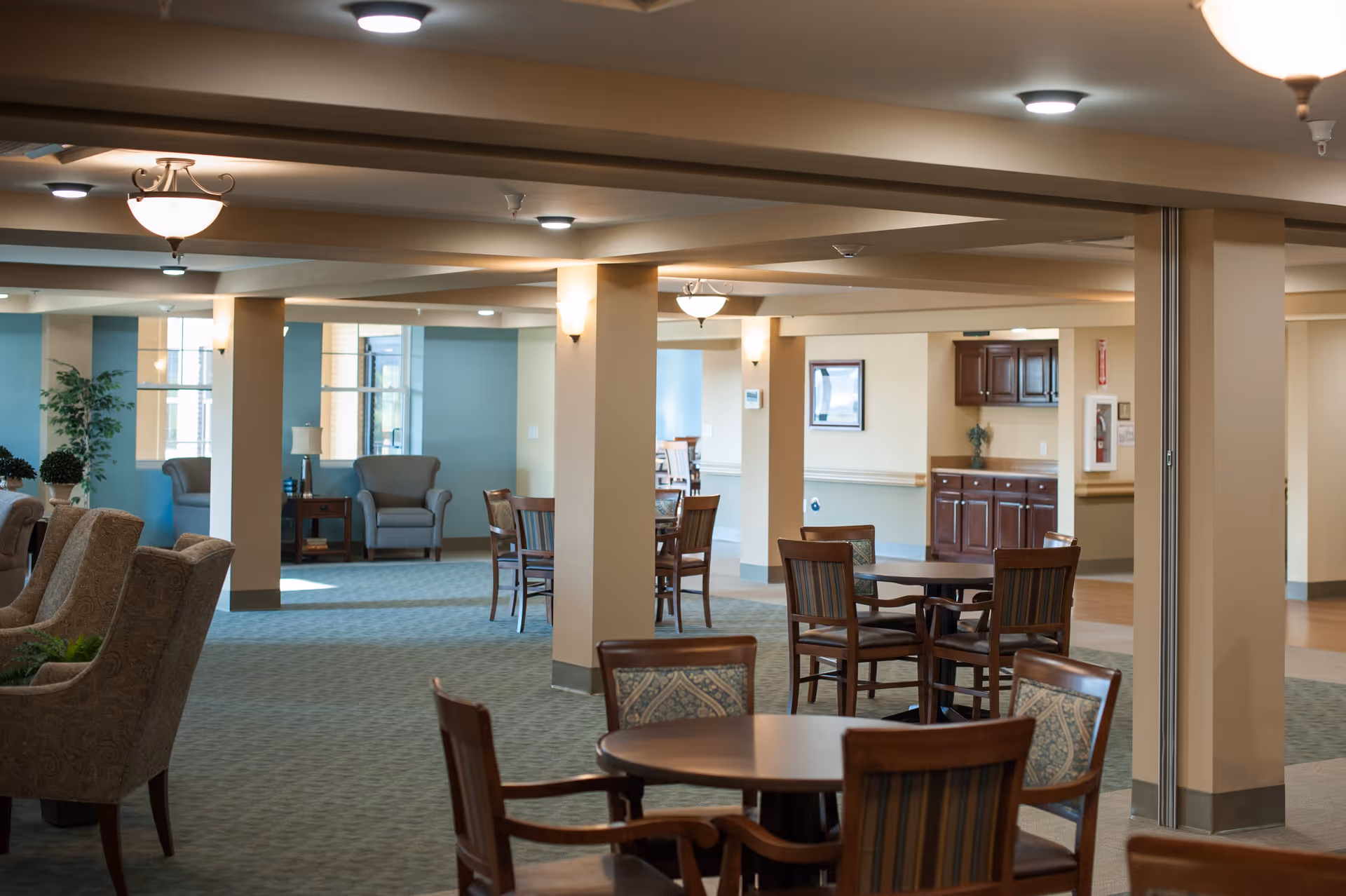 Spacious senior living common area with multiple round wooden tables and chairs, beige pillars, soft lighting fixtures on the ceiling, and comfortable armchairs near windows with natural light.