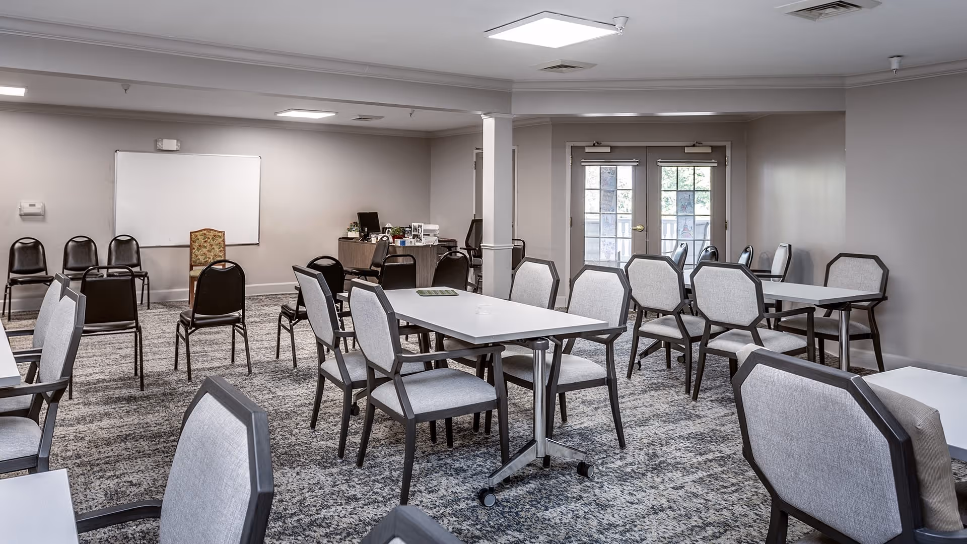 A spacious room with multiple tables and chairs arranged for group seating. The room has a carpeted floor, neutral-colored walls, and a whiteboard on one wall. There is a desk with office supplies and a computer in the corner, and double glass doors leading outside.