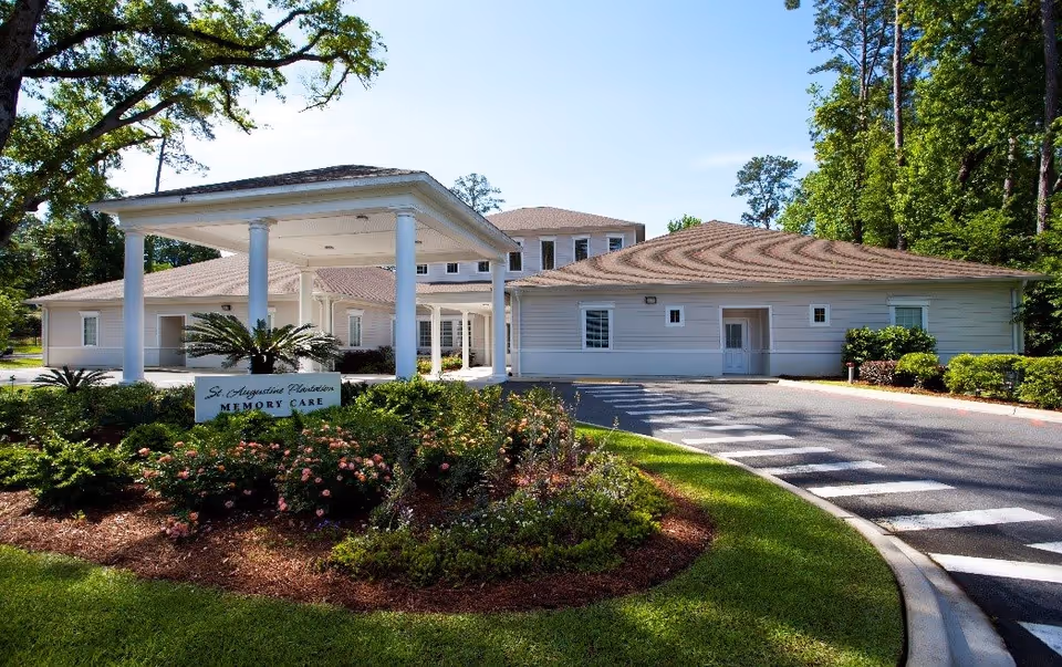 Exterior view of St. Augustine Plantation Memory Care facility showing a single-story building with a covered entrance supported by white columns, surrounded by landscaped greenery and flowers under a clear blue sky.