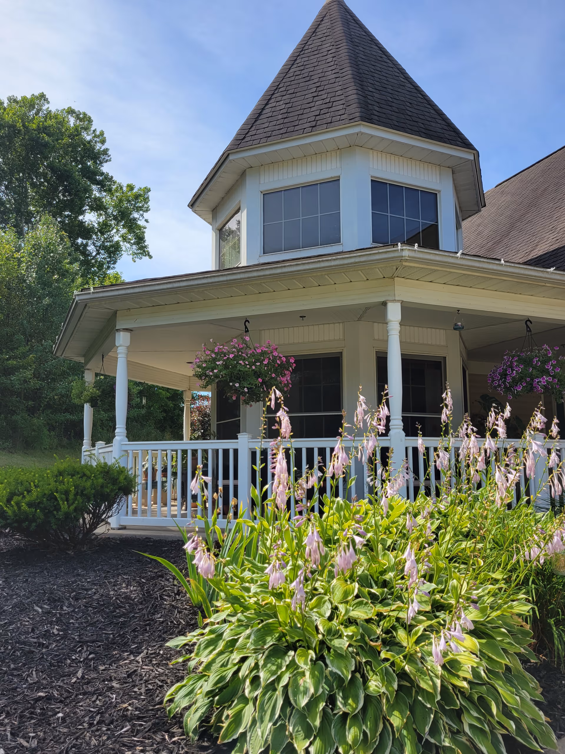 Exterior view of a building with a pointed roof and large windows, featuring a white porch with railings and hanging flower baskets. In the foreground, there are green plants with light purple flowers and a mulched garden bed. Trees and a clear blue sky are visible in the background.