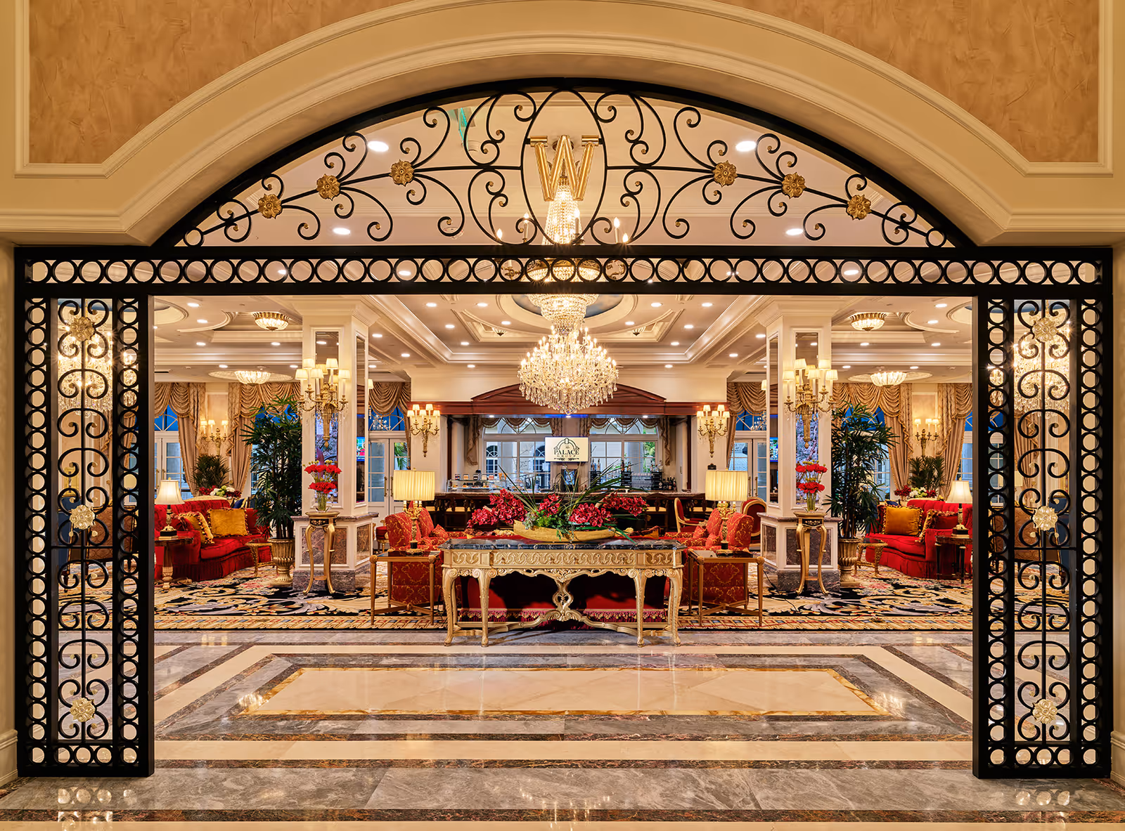 Ornate wrought-iron arched entry with a gold 'W' opening into an opulent lobby featuring chandeliers, red seating, and decorative tables.