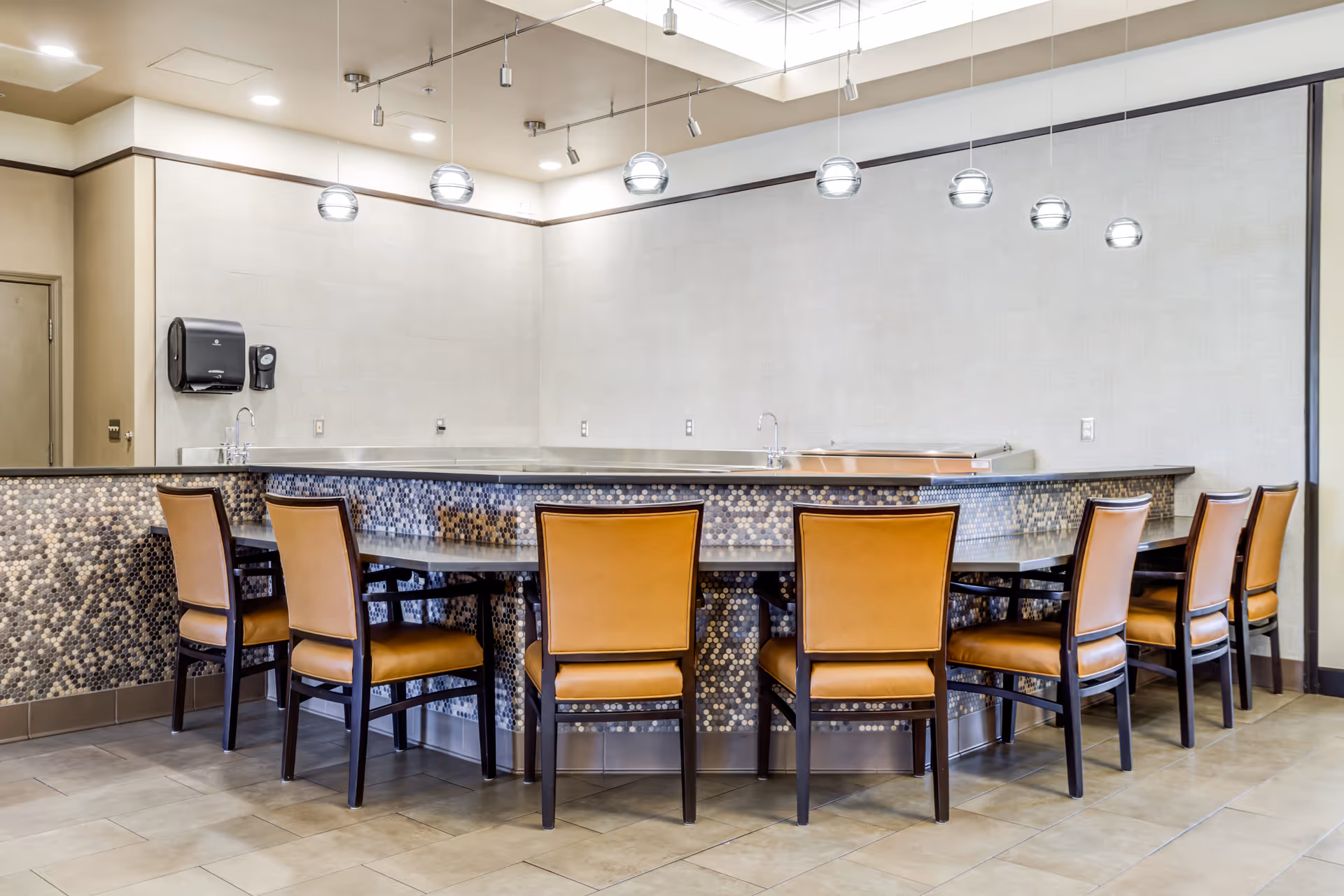 A modern dining area with a curved counter featuring a mosaic tile front and eight brown cushioned chairs arranged around it. Above the counter, there are seven spherical pendant lights hanging from the ceiling. The walls are light-colored and there is a paper towel dispenser and soap dispenser mounted on the wall near a door in the corner.