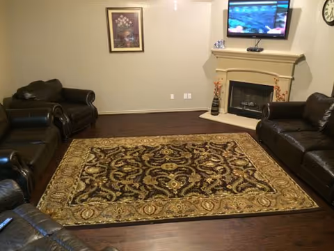 Living room with dark leather sofas arranged around a large patterned area rug, a TV mounted above a fireplace, and framed art on the wall.