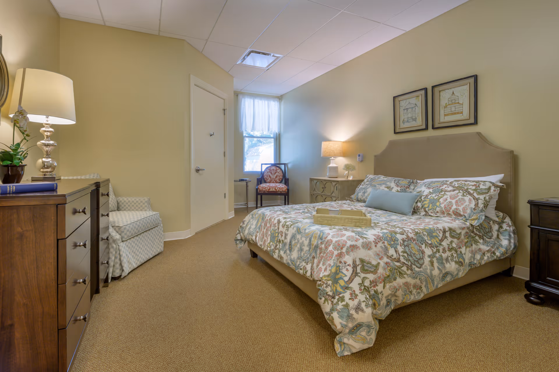 A cozy bedroom with a large bed covered in a floral patterned comforter and multiple pillows. There is a wooden dresser with a lamp and a plant on it to the left, a cushioned armchair next to the dresser, and a small wooden chair near a window with sheer curtains. On the right side of the bed is a nightstand with a lamp and decorative items. The walls are painted a soft beige color and two framed pictures hang above the bed.