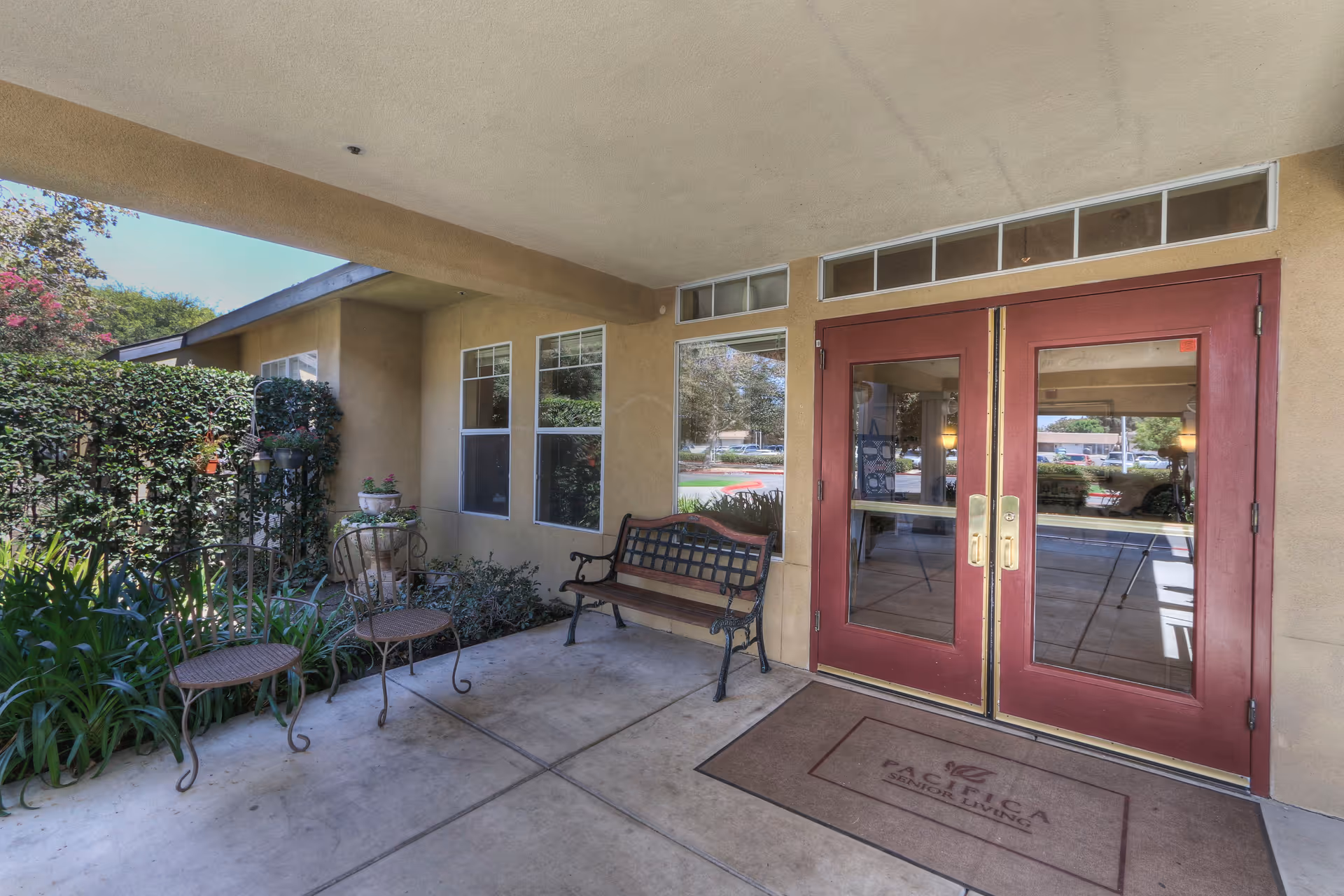 Covered entrance area of a senior living facility with a bench, two small round tables, and two chairs. The entrance has double red doors with glass panels and a doormat that reads 'Pacifica Senior Living'. There are plants and greenery along the side wall and windows.