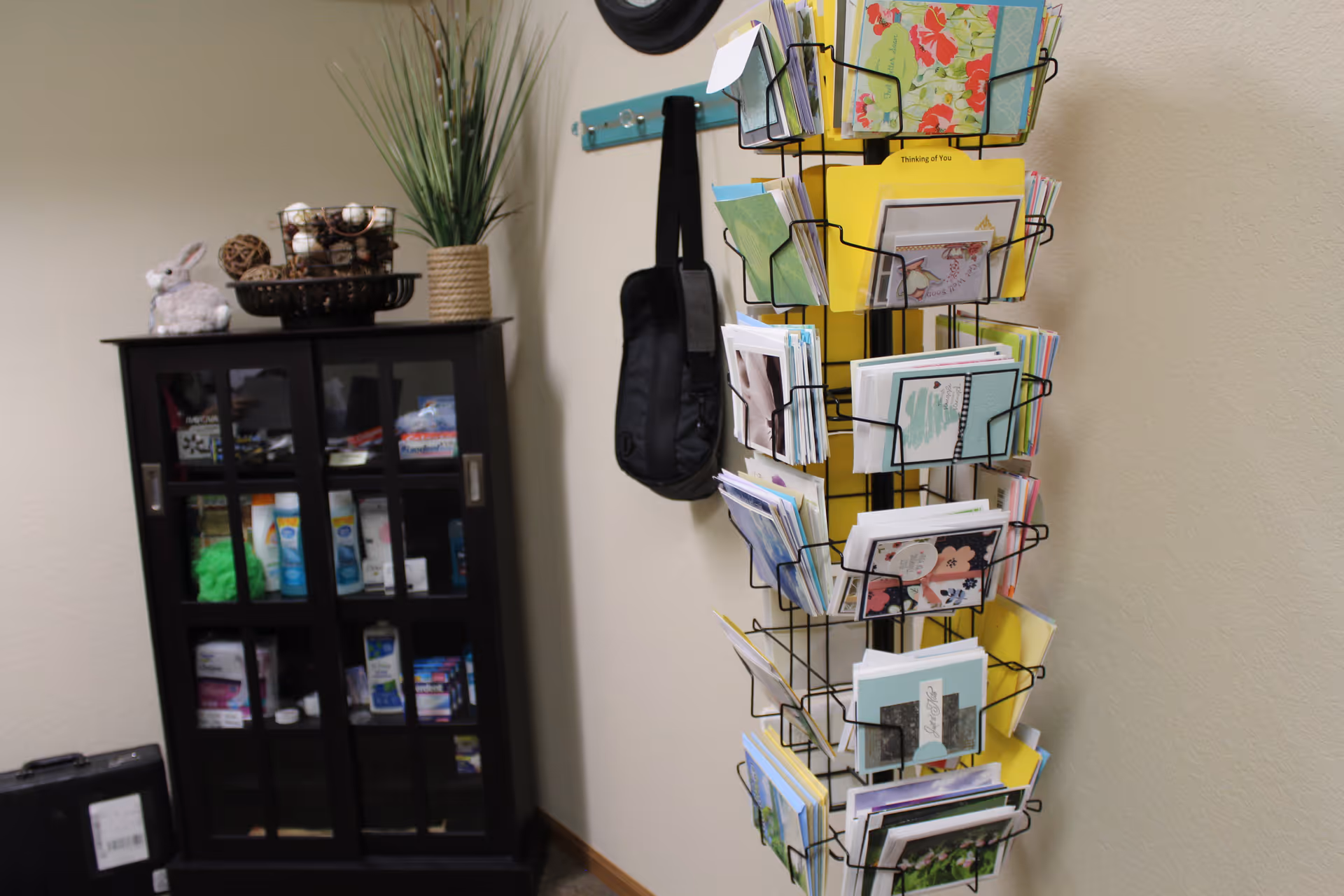 A wall-mounted rotating rack filled with greeting cards next to a small glass-front cabinet and decorative items in an interior space.