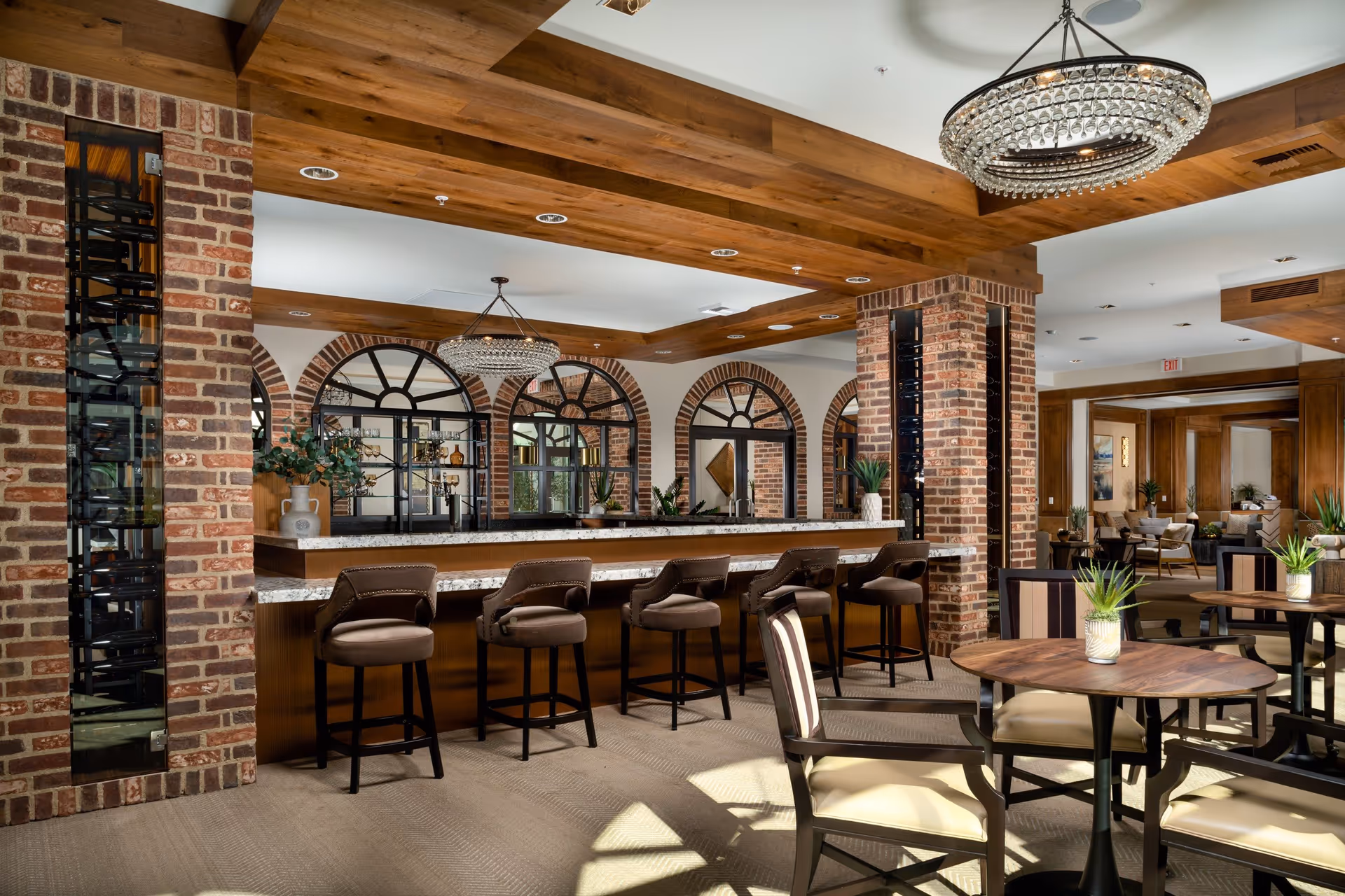 A spacious and elegant common area with a long marble countertop bar featuring six cushioned bar stools. The room has exposed wooden beams on the ceiling, brick pillars, and large arched windows with black frames. There are round wooden tables with cushioned chairs and small potted plants on the tables. A large crystal chandelier hangs from the ceiling, and the area extends into a lounge with additional seating and plants.
