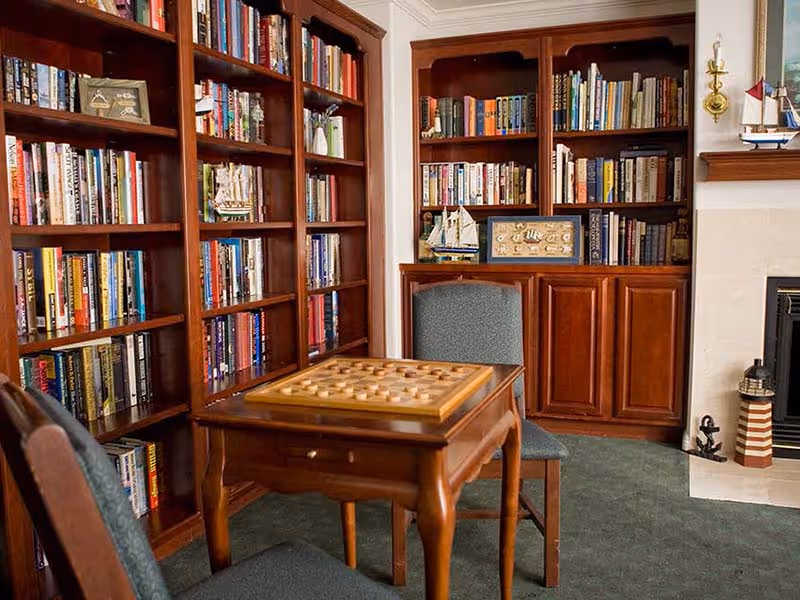 Cozy library room with wooden bookshelves filled with books, a wooden table with a checkers board on top, two upholstered chairs, and a fireplace with nautical-themed decorations including model ships and a lighthouse figurine.