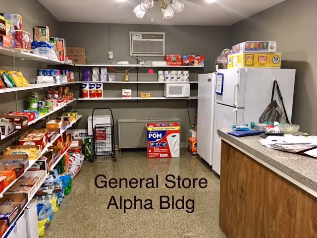 Interior of a small general store room with shelves of snacks and supplies, a refrigerator, microwave, and a countertop labeled 'General Store Alpha Bldg'.