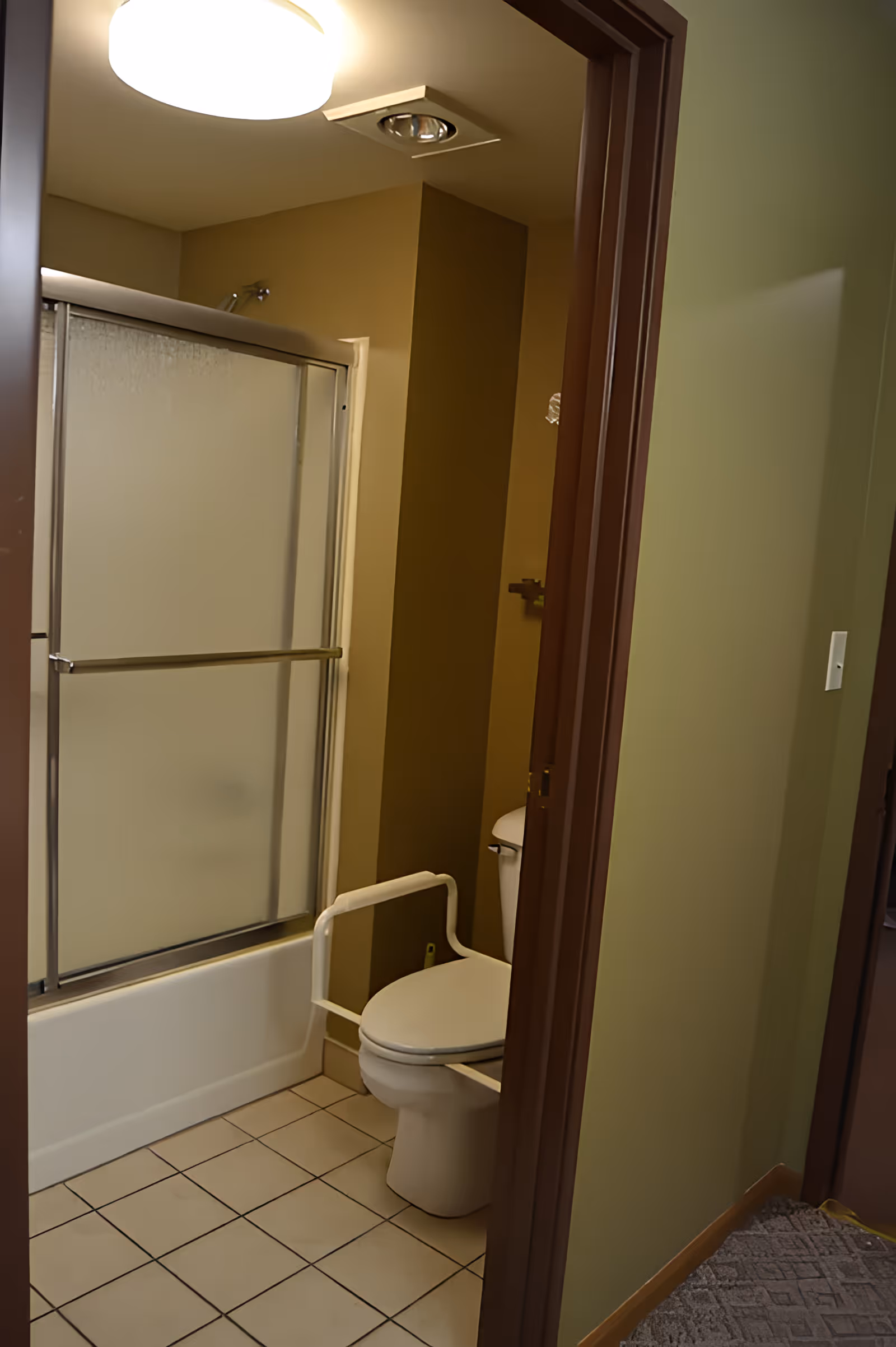 Bathroom seen from the doorway with a toilet, a safety grab bar, and a bathtub with frosted sliding shower doors.