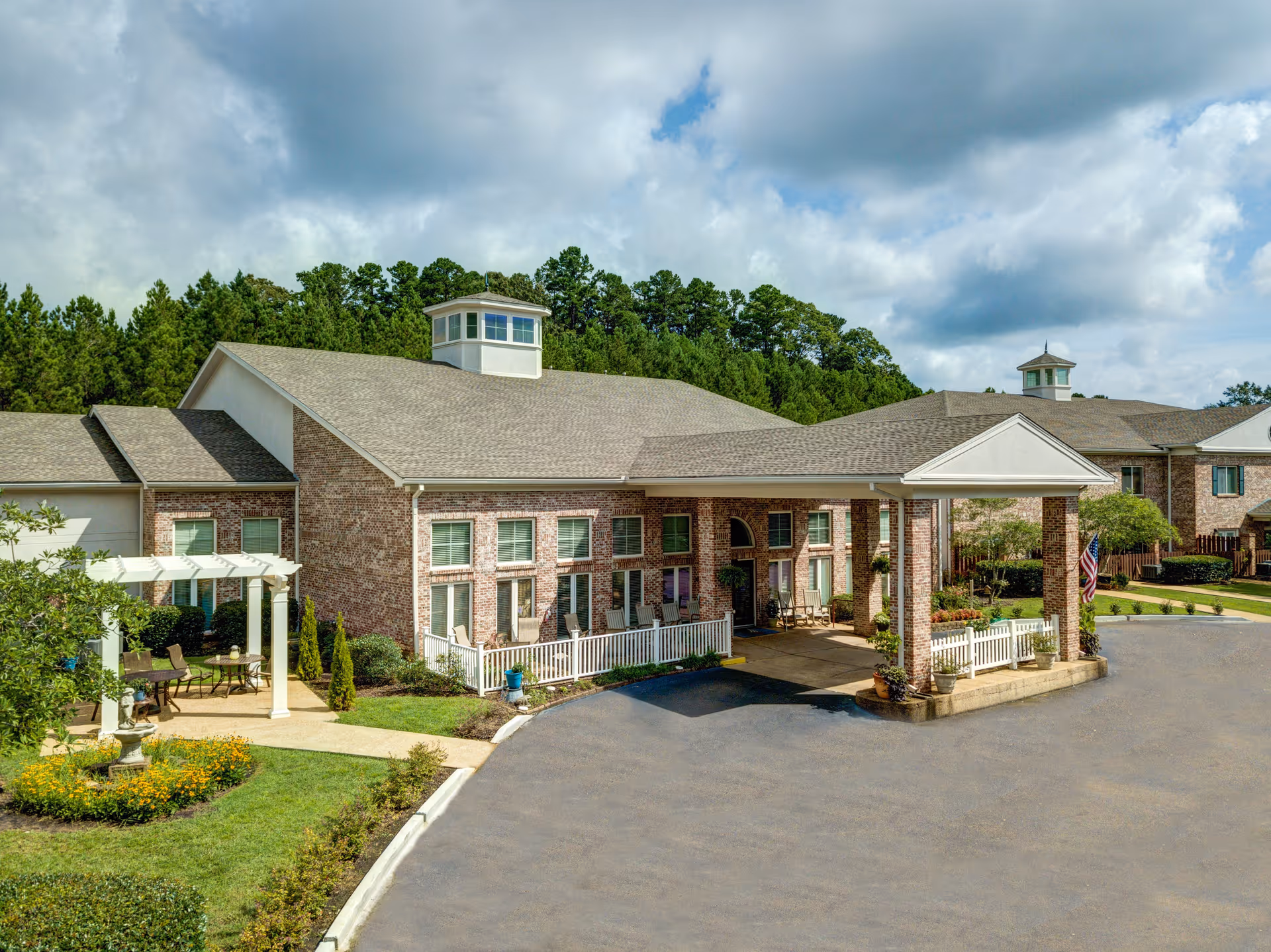 Exterior view of a senior living facility named Castlewoods Place featuring a brick building with a covered entrance, multiple windows, a small white fence, outdoor seating area with chairs and tables, landscaped garden with flowers, and a backdrop of green trees under a partly cloudy sky.
