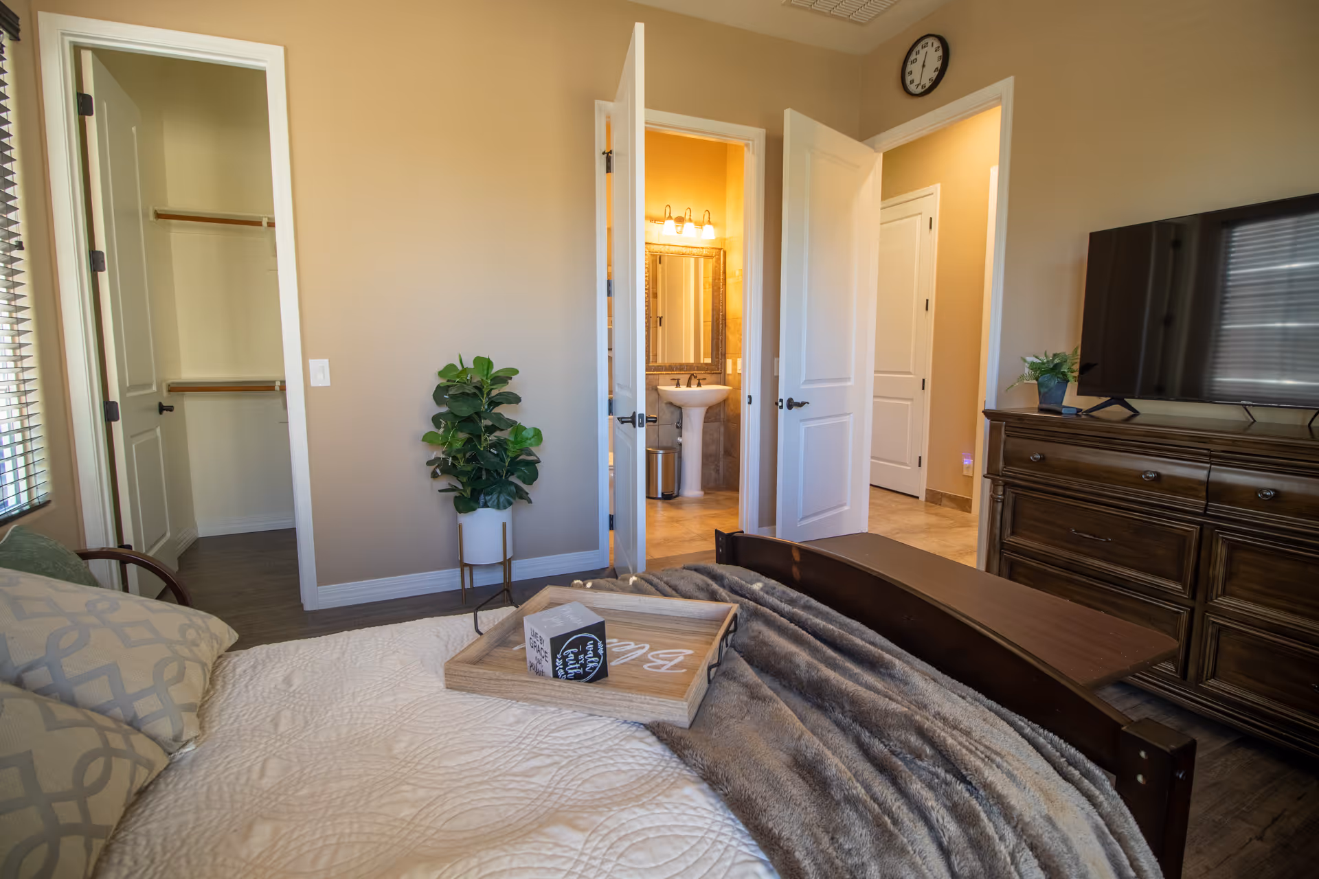 A cozy bedroom in an assisted living facility with a bed covered in a white quilt and gray blanket, decorative pillows, a wooden tray with decorative blocks on the bed, a wooden dresser with a flat-screen TV, a potted plant on the dresser, an open door leading to a bathroom with a pedestal sink and mirror, and another open door leading to a closet.