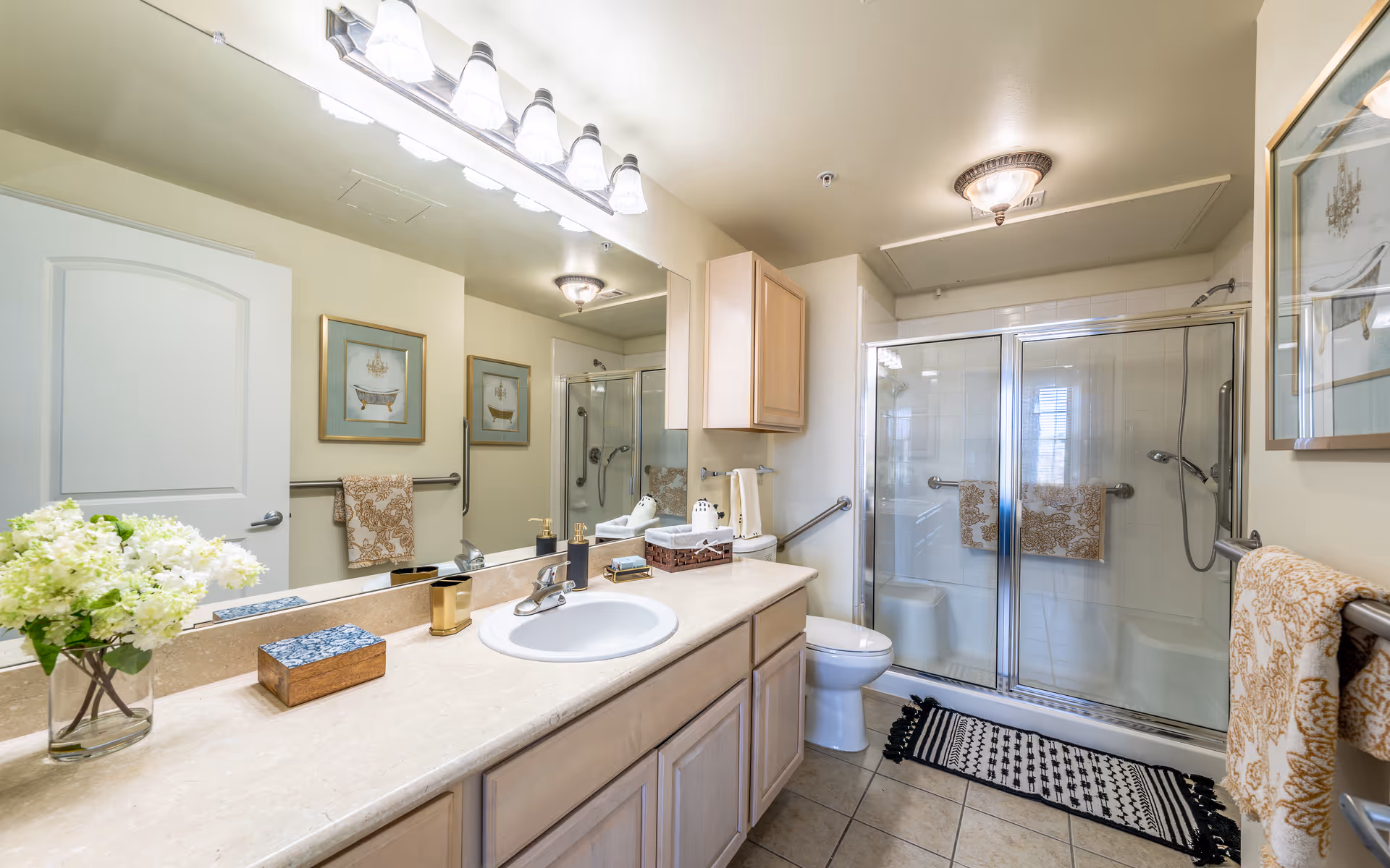 A clean and well-lit bathroom featuring a large mirror above a beige countertop with a sink. On the countertop are a vase with white flowers, a soap dispenser, a tissue box, and other small items. The bathroom has a toilet next to a glass-enclosed shower with sliding doors. There are two framed pictures on the walls, patterned towels hanging on rails, and a decorative rug on the tiled floor.