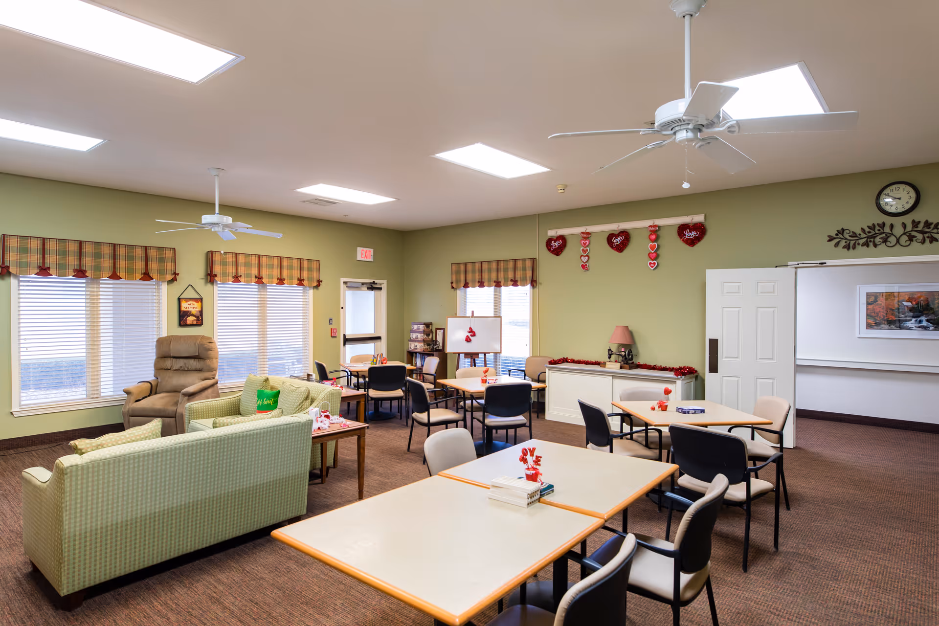 A well-lit common room in an assisted living facility with several tables and chairs arranged for group activities or dining. The room features green walls, plaid valances on the windows, ceiling fans, a recliner chair, and a green couch. Valentine's Day decorations, including hanging hearts and table centerpieces, add a festive touch. A clock and framed artwork are visible on the walls.