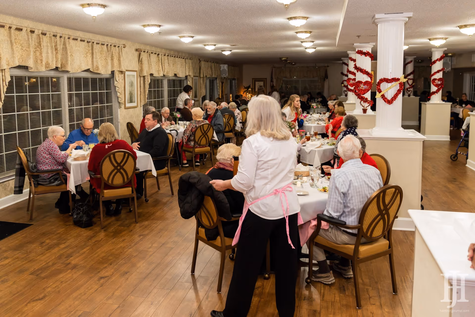 A dining room in a senior living facility with multiple elderly residents seated at tables enjoying a meal. A staff member wearing a white shirt and pink apron is standing and attending to the guests. The room has wooden floors, large windows with curtains, and white columns decorated with red heart-shaped garlands.