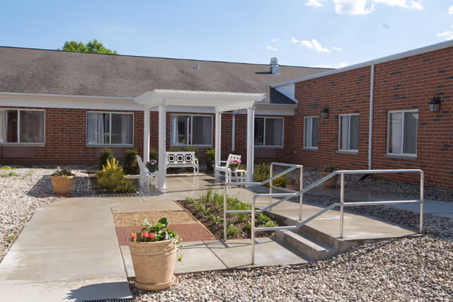 Outdoor courtyard area of a senior living facility with a white pergola covering a bench and chairs, surrounded by brick building walls and windows. There are paved walkways, a ramp with handrails, and potted plants with flowers. The sky is clear with a few clouds.