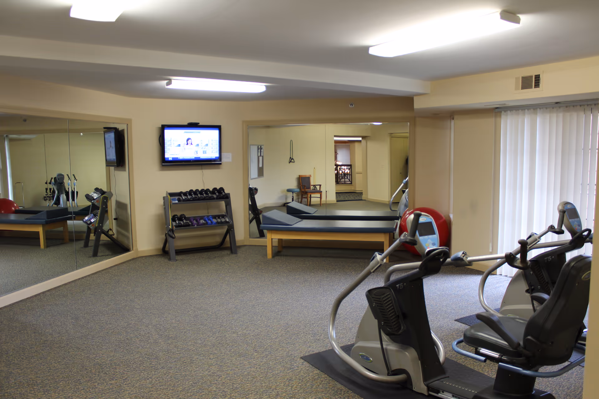 A senior facility exercise room with cardio machines, a weight rack, mirrors, and a wall-mounted TV.
