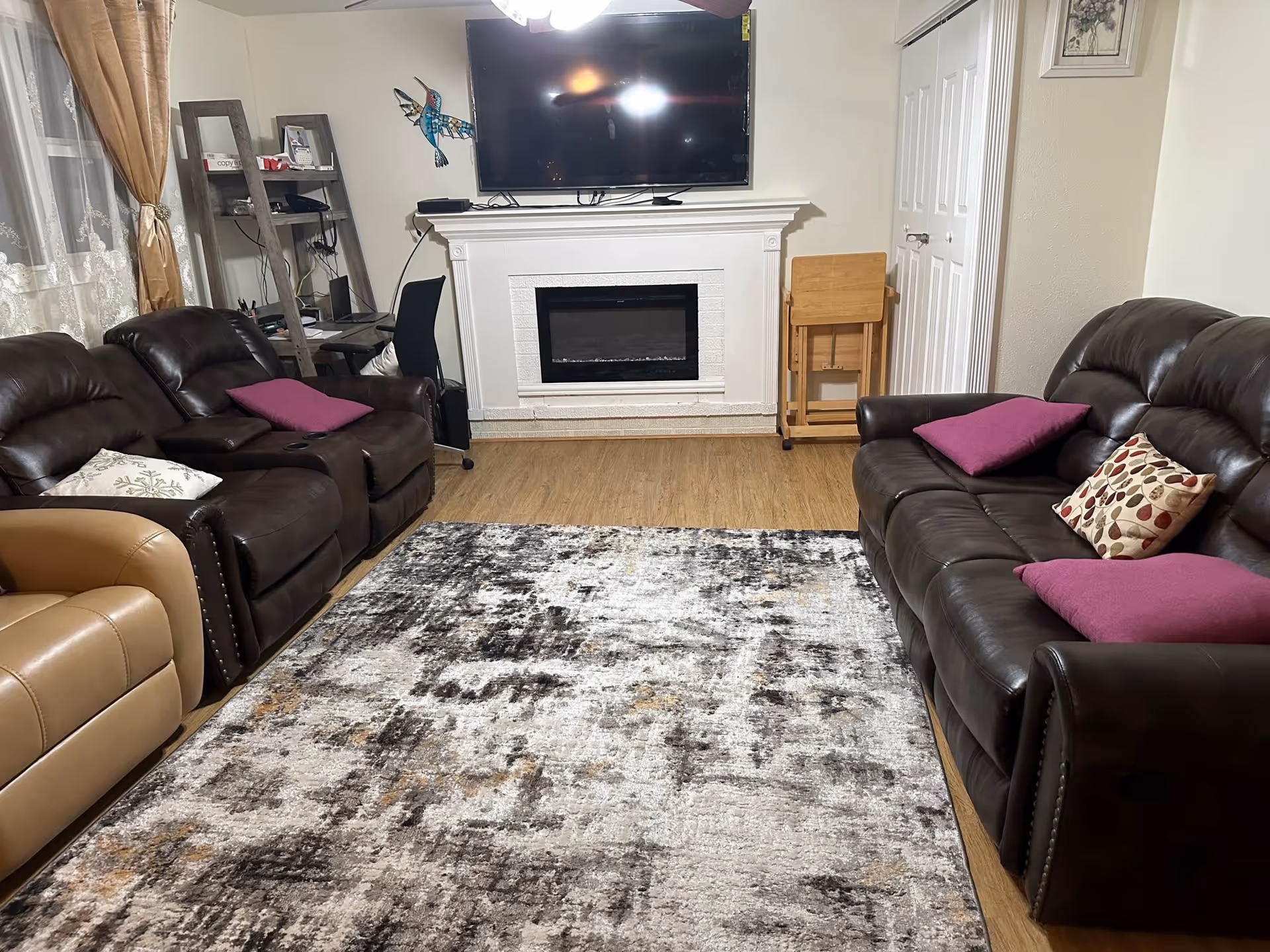 Cozy living room with two leather sofas facing a patterned rug, a wall-mounted TV above a white fireplace, and decorative pillows.