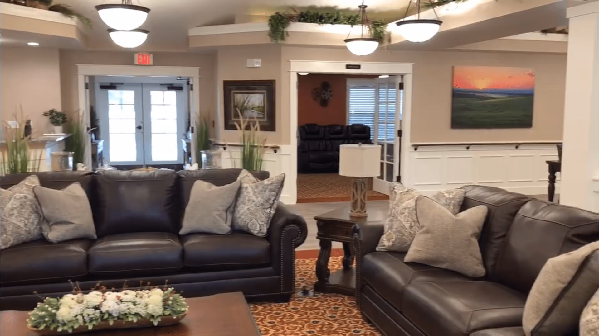 A cozy assisted living facility common area with two dark leather sofas adorned with multiple patterned and plain cushions. A wooden coffee table with a floral centerpiece is in front of the sofas. The room features warm lighting, a patterned carpet, and decorative plants. In the background, there is a doorway leading to another room with additional seating and a large window with blinds. The walls are decorated with framed artwork and wainscoting.