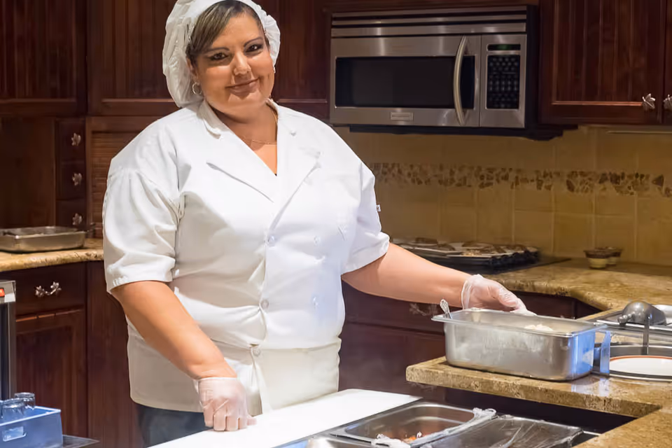 A chef wearing a white uniform and hairnet stands in a kitchen, serving food from a metal container on a countertop. The kitchen has dark wooden cabinets, a microwave, and a stove in the background.